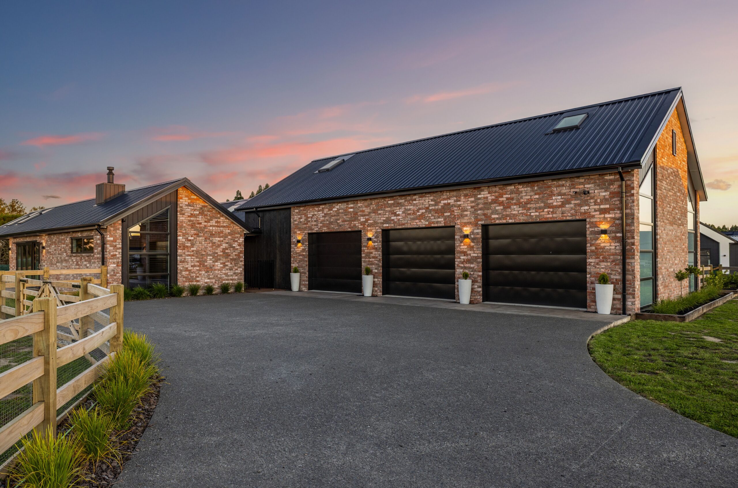 Hallmark Homes brick house exterior at sunset, featuring a triple garage with black panelled doors, aged brick cladding, black corrugated iron roof, warm wall-mounted sconce lighting, and a sweeping asphalt driveway bordered by a timber post-and-rail fence.