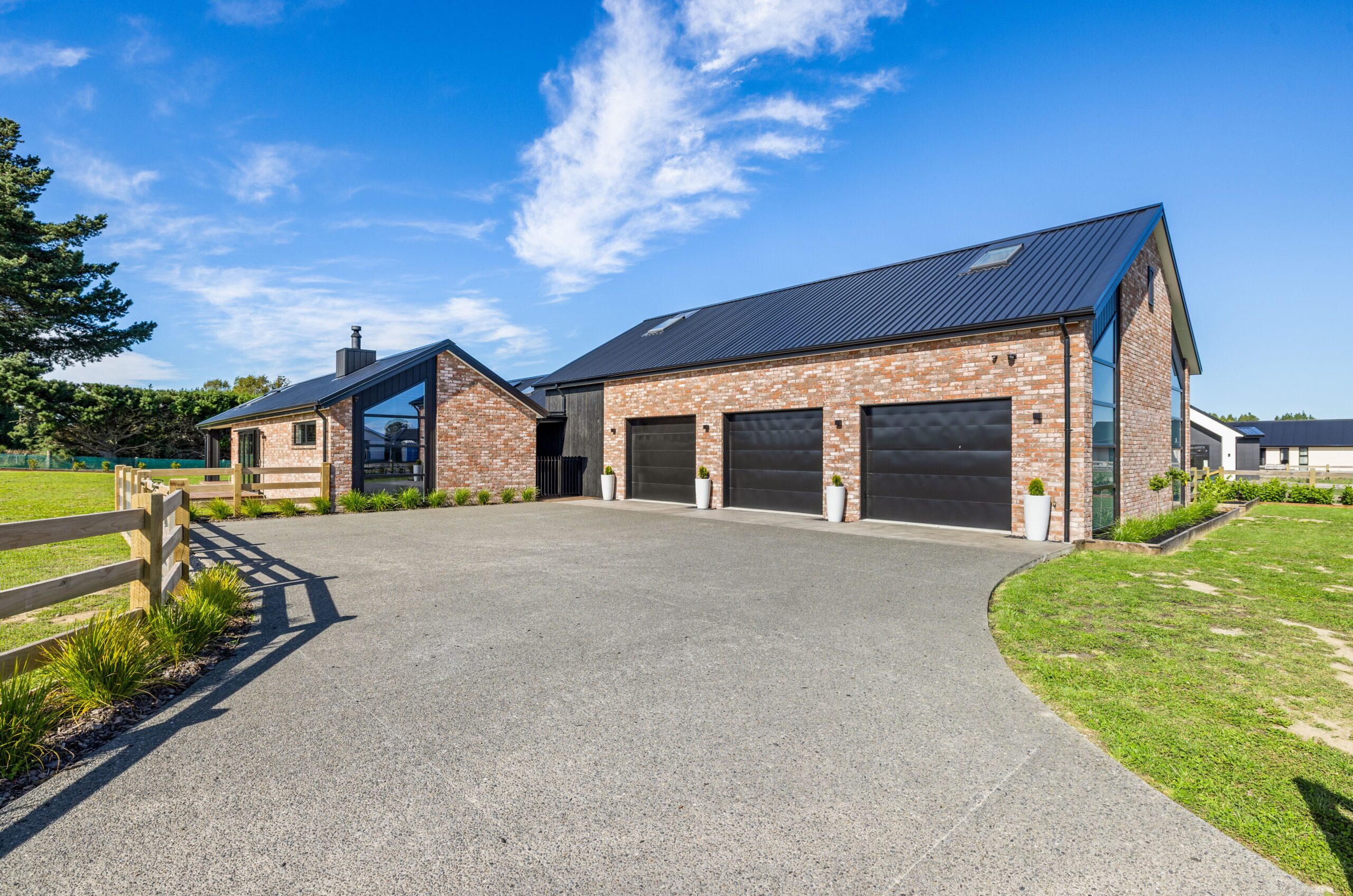 Daytime front elevation of a Hallmark Homes brick build showing triple black garage doors, black corrugated iron roof, dark-stained timber feature panel, floor-to-ceiling glazing on the gable end, and a curved asphalt driveway with lush grass surrounds.