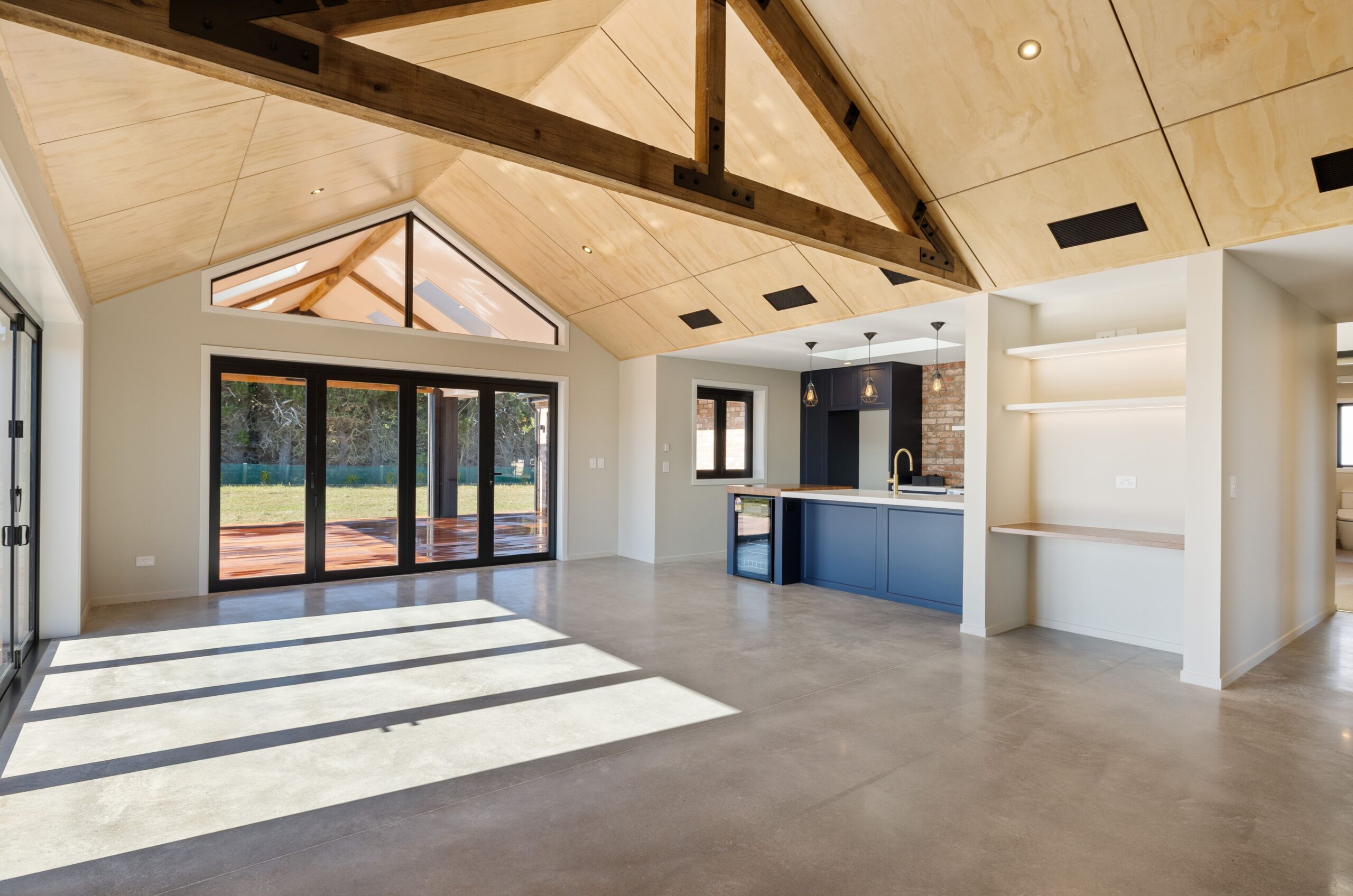 Open plan living and kitchen in a Hallmark Homes residence showing a plywood vaulted ceiling with dark exposed king post trusses, a triangular glazed gable window, polished concrete floors, black bi-fold doors to the deck, navy blue kitchen island with cage pendant lighting, and built-in shelving.