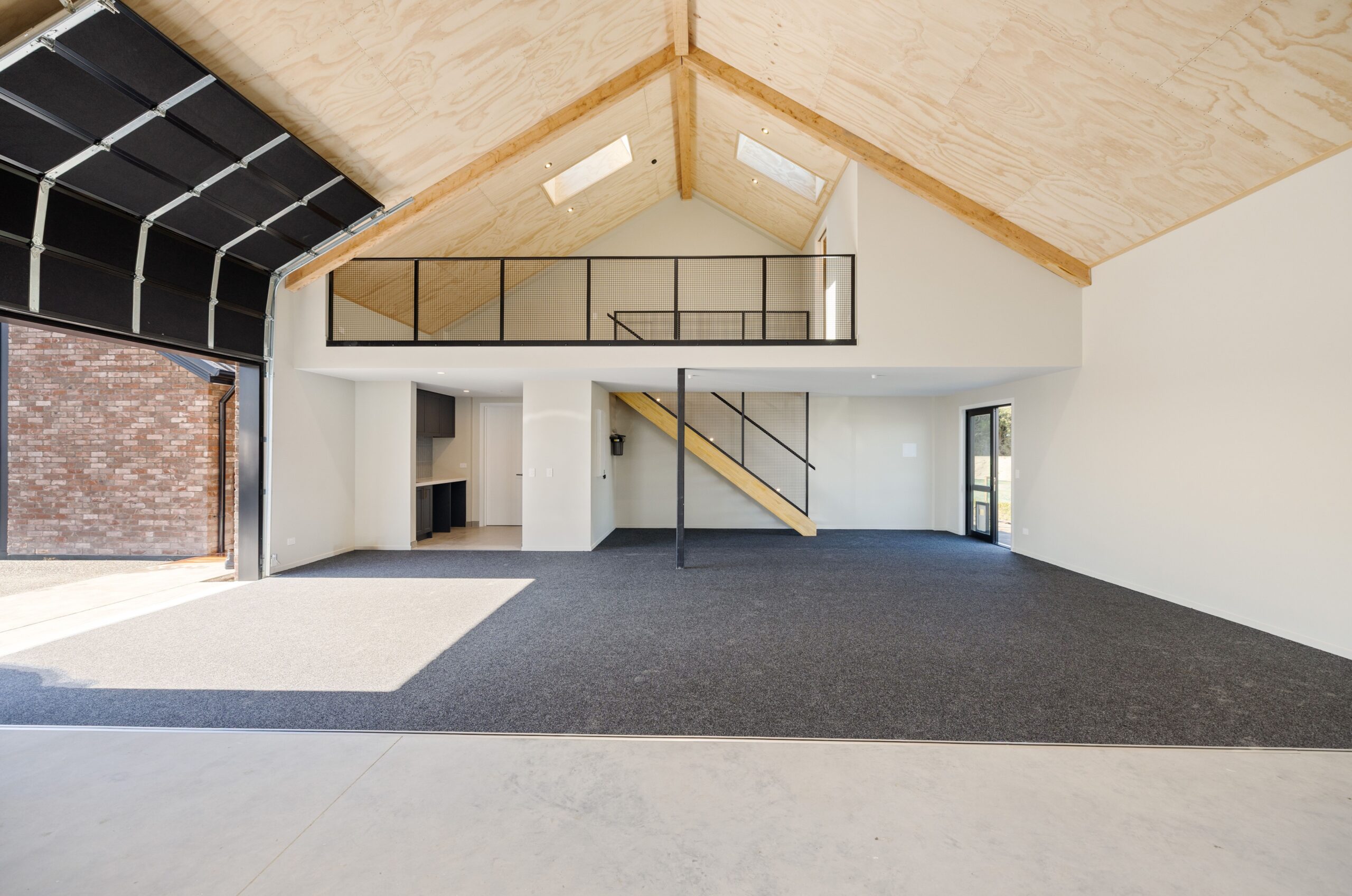 Interior of a Hallmark Homes lifestyle shed featuring a soaring plywood cathedral ceiling with exposed timber rafters, a mezzanine level with black steel mesh balustrade, timber staircase, built-in kitchenette, and polished concrete floor transitioning to carpet.