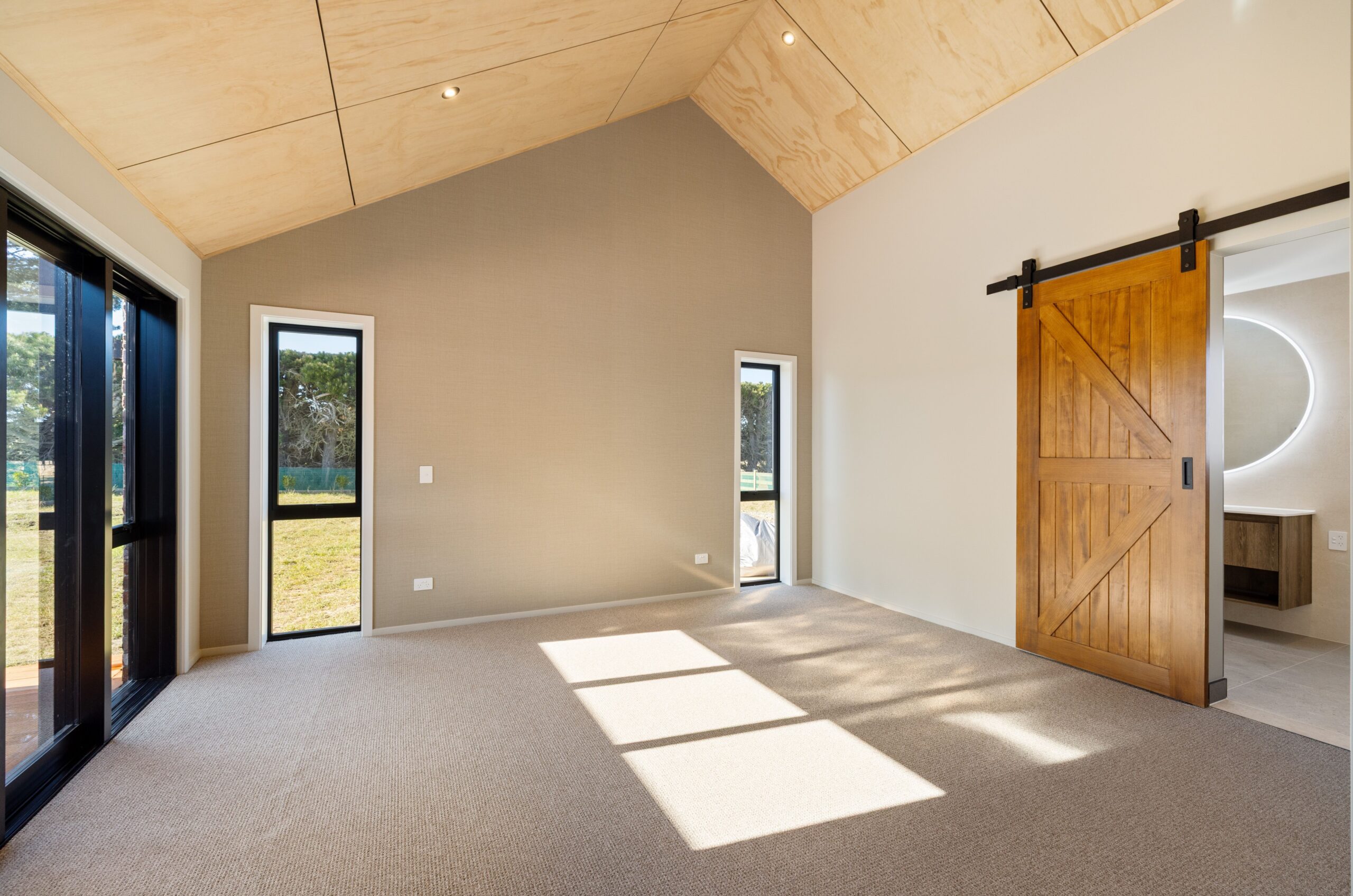 Spacious master bedroom with a plywood vaulted ceiling, carpet flooring, neutral warm-toned walls, black-framed windows overlooking the garden, and a sliding timber barn door on a black track opening to an ensuite with a round LED mirror visible beyond.