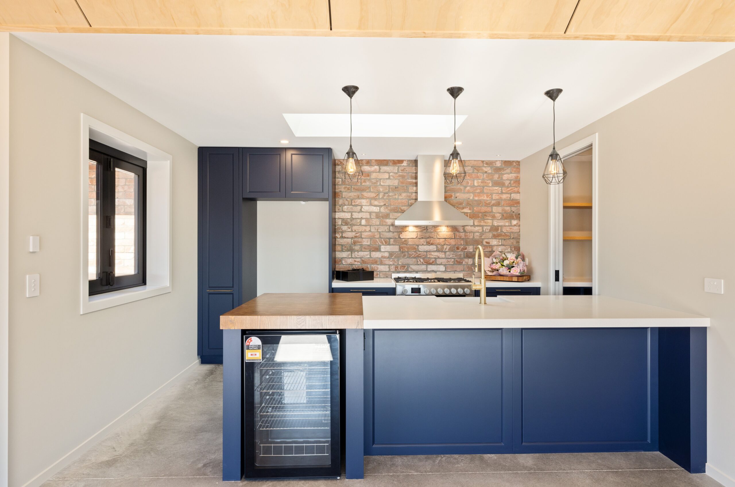 Detail shot of a Hallmark Homes kitchen showing navy blue shaker cabinetry with brushed gold handles, a white quartz benchtop, a Bosch freestanding gas range with stainless steel rangehood, an exposed brick splashback, and an open butler's pantry with timber shelving to the right.