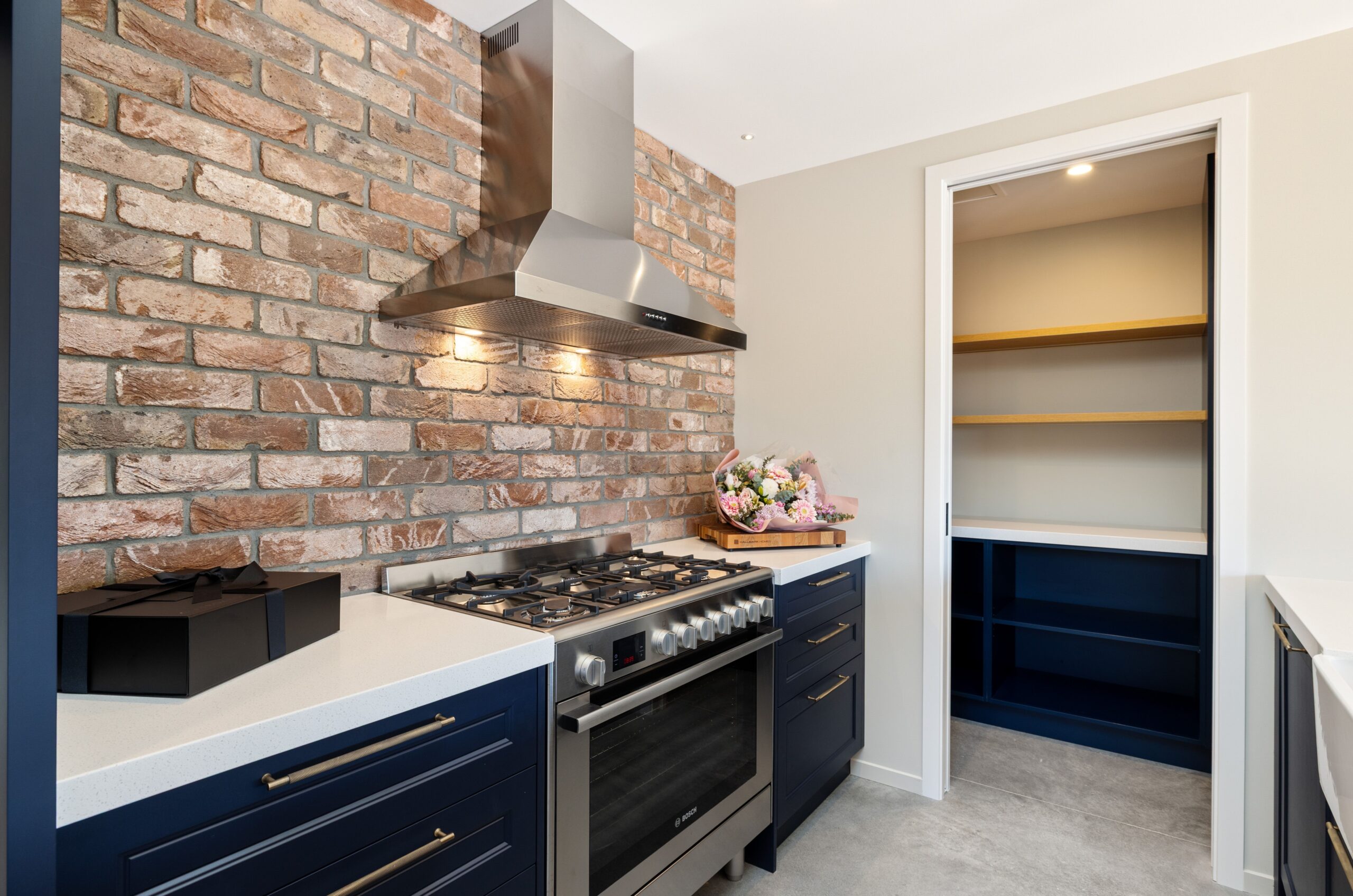 Wide kitchen view featuring a navy blue island with a white quartz waterfall top, timber butcher block breakfast bar section, under-bench wine fridge, black cage pendant lights overhead, an exposed brick feature wall behind the cooktop, and a skylight flooding the space with natural light.