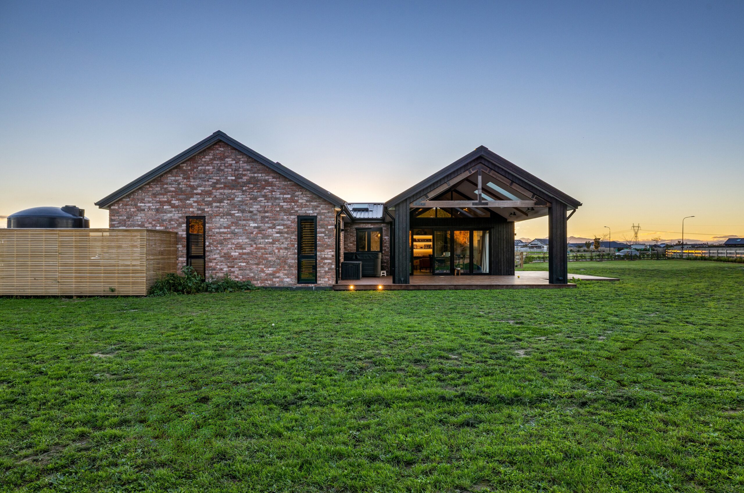 Rear view of a Hallmark Homes brick build at dusk, showcasing a dark-stained timber gable pergola with exposed beam structure, open-plan indoor/outdoor living space, warm interior lighting glowing through large glazed doors, and an expansive lawn.