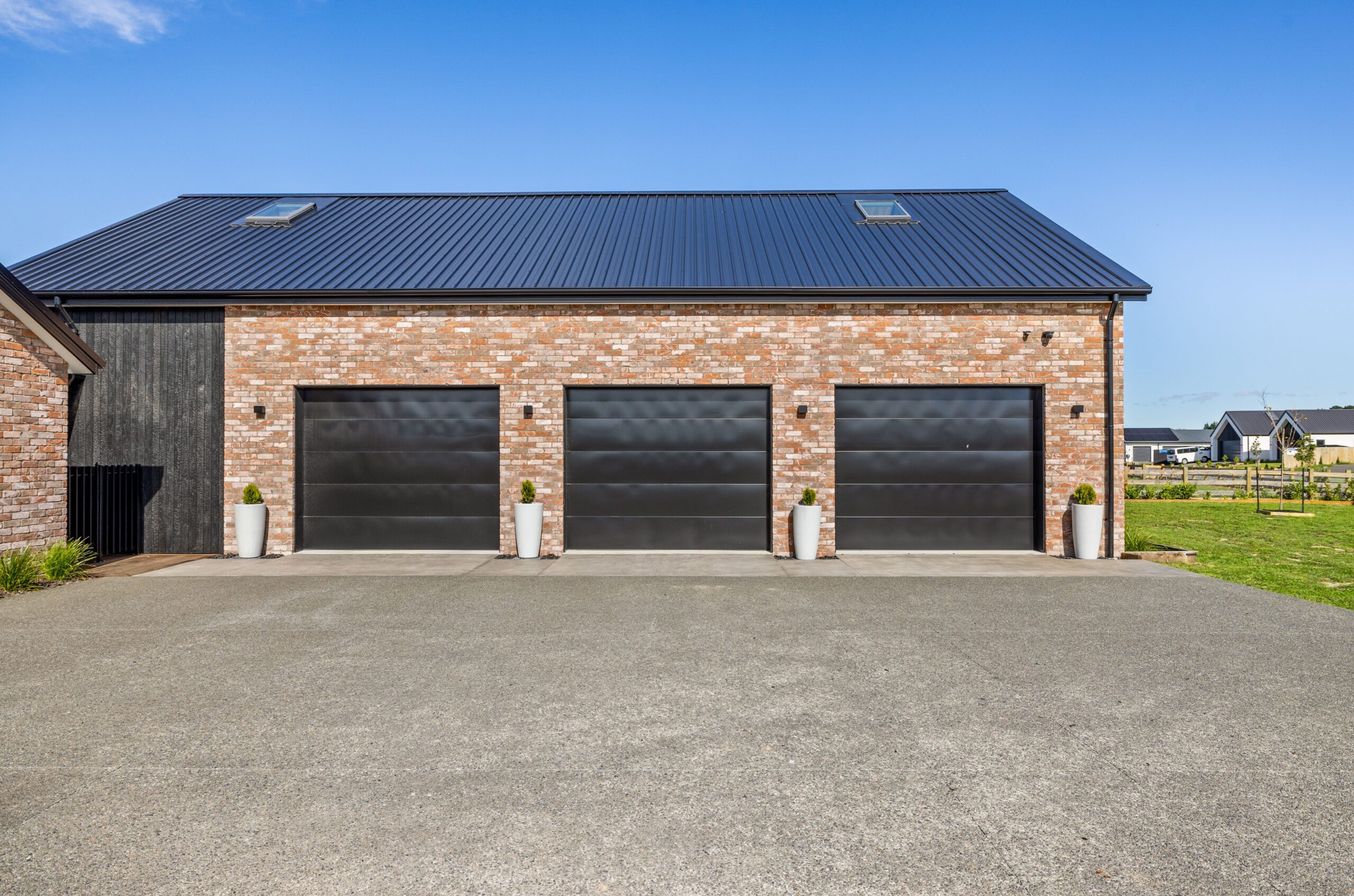 Close-up of a Hallmark Homes triple garage facade with three large black sectional doors set into aged brick cladding, black corrugated iron roof, wall-mounted security lights, and white cylindrical planters flanking each bay.