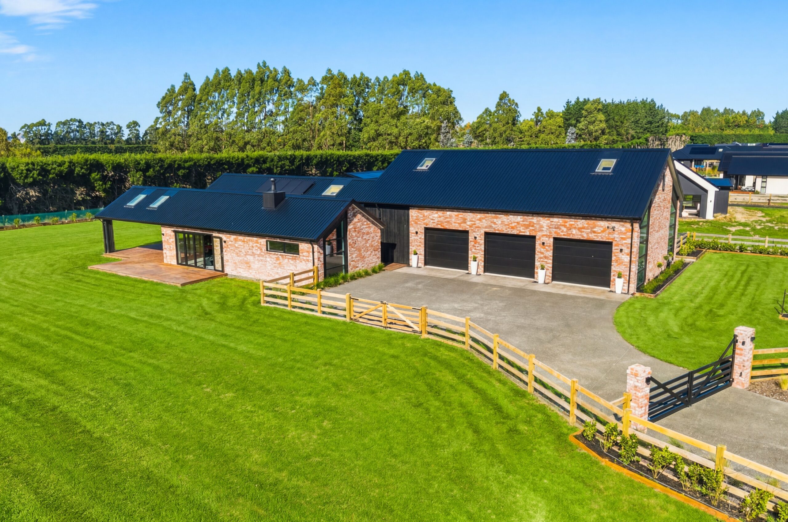 Aerial view of a Hallmark Homes lifestyle property showing the full brick and black-roofed residence with attached shed, triple garage, timber deck, wide concrete driveway, timber post-and-rail paddock fencing, and manicured lawns backed by mature trees.