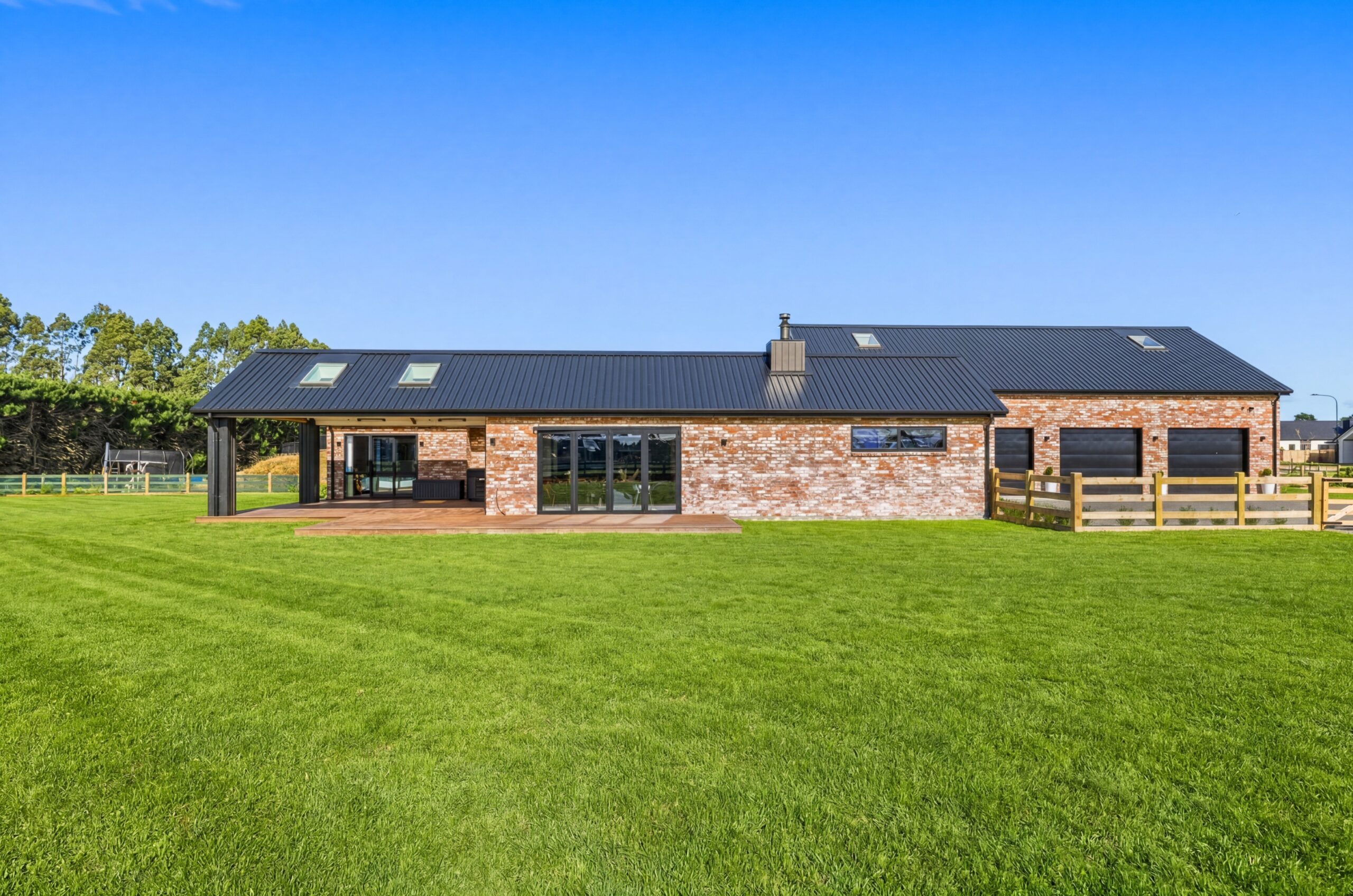Rear exterior of a long-form Hallmark Homes brick residence under a clear blue sky, with a black metal roof, skylights, bi-fold aluminium doors opening onto a timber deck, and a large manicured lawn with a timber paddock fence to the right.