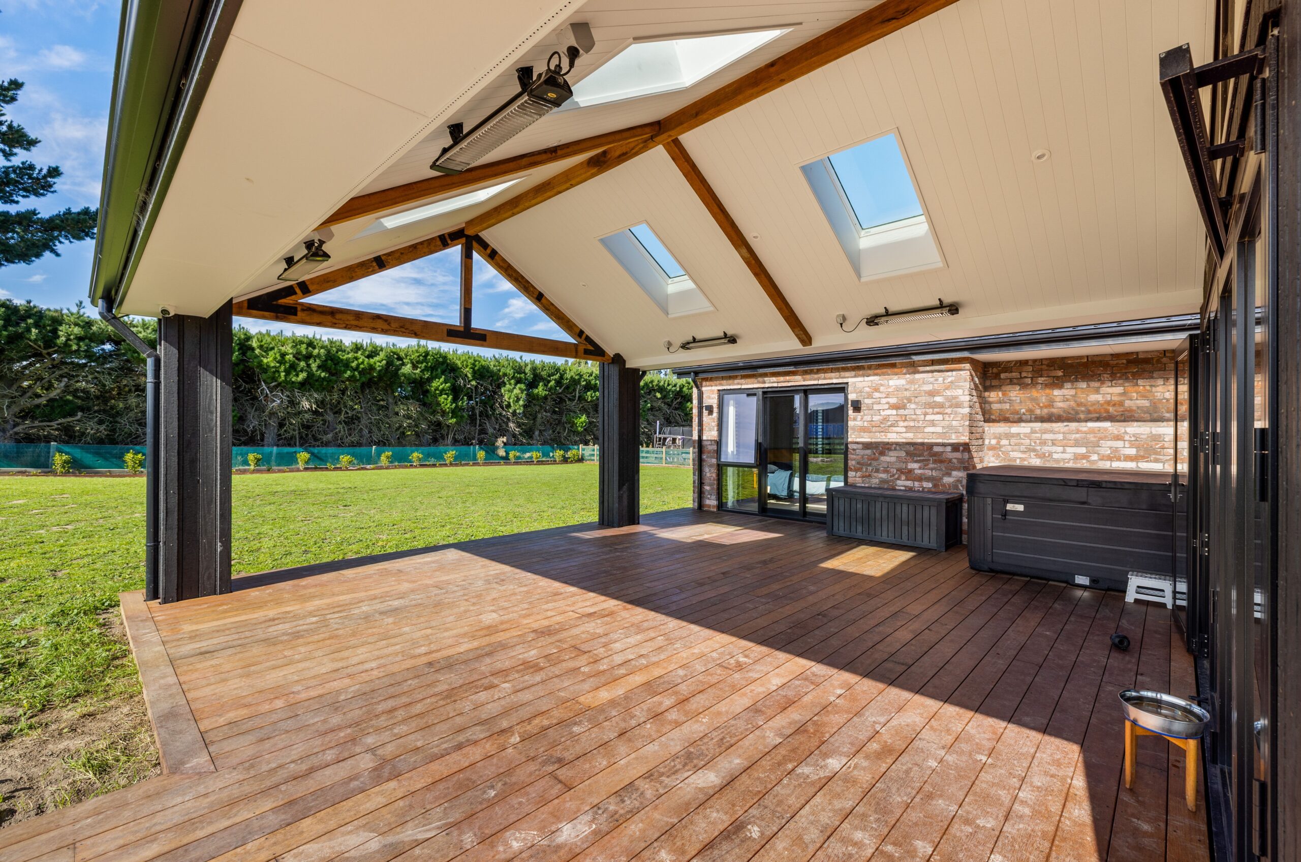 Spacious covered alfresco deck with warm timber decking, an exposed-beam gable roof with multiple skylights and infrared heaters, dark-stained timber posts and fascia, a hot tub in the corner, and views across an open lawn through the open sides.