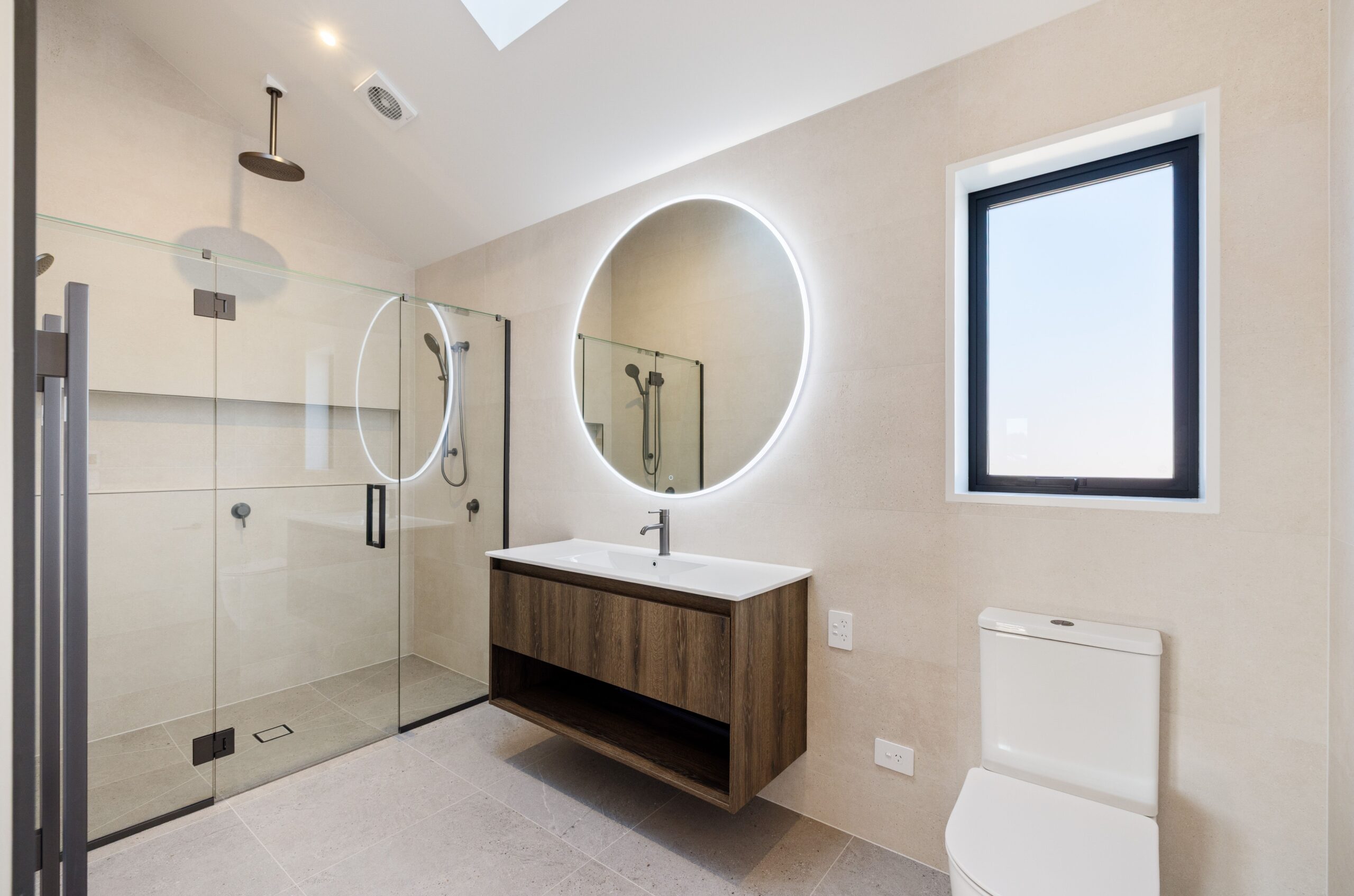 Modern bathroom featuring a frameless glass walk-in shower with matte black fittings and a ceiling-mounted rain head, a wall-hung walnut timber vanity with white basin, a large round LED backlit mirror, stone-look floor tiles, and a skylight above.