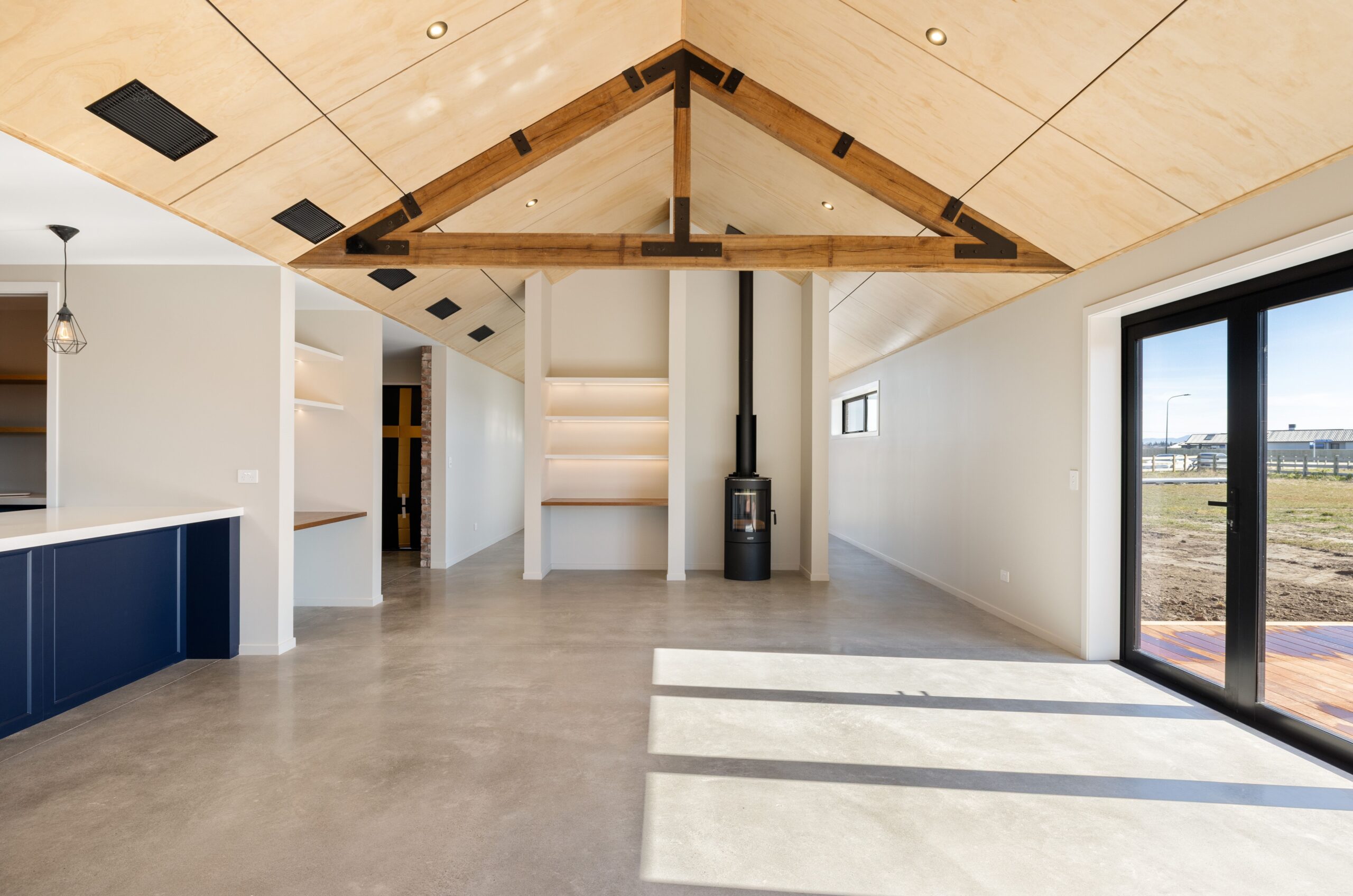 Bright open-plan living area in a Hallmark Homes build with polished concrete floors, a soaring plywood cathedral ceiling featuring exposed dark timber king post trusses, a freestanding black wood burner, navy blue kitchen cabinetry visible to the left, and bi-fold doors opening to a timber deck.