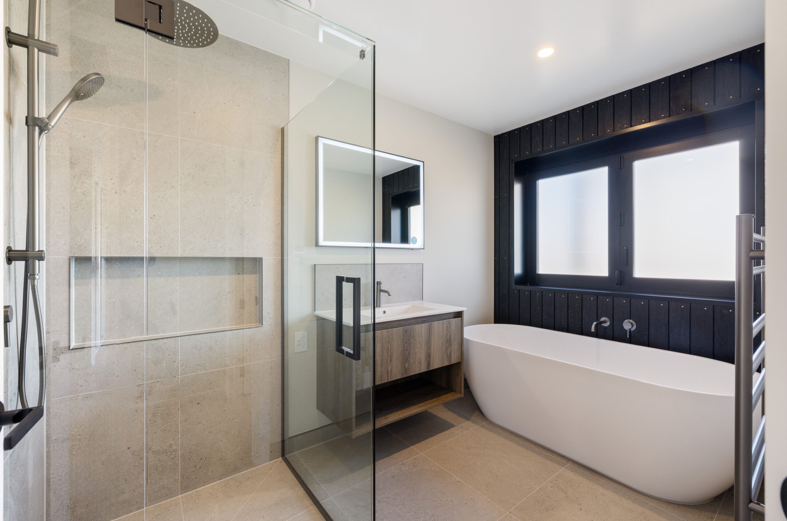 Luxurious Bathroom with a large frameless glass shower enclosing stone-look tiled walls and a built-in niche, a freestanding white soaker bath, a wall-hung timber vanity with rectangular LED mirror, dark vertical timber feature wall, and a chrome heated towel rail.