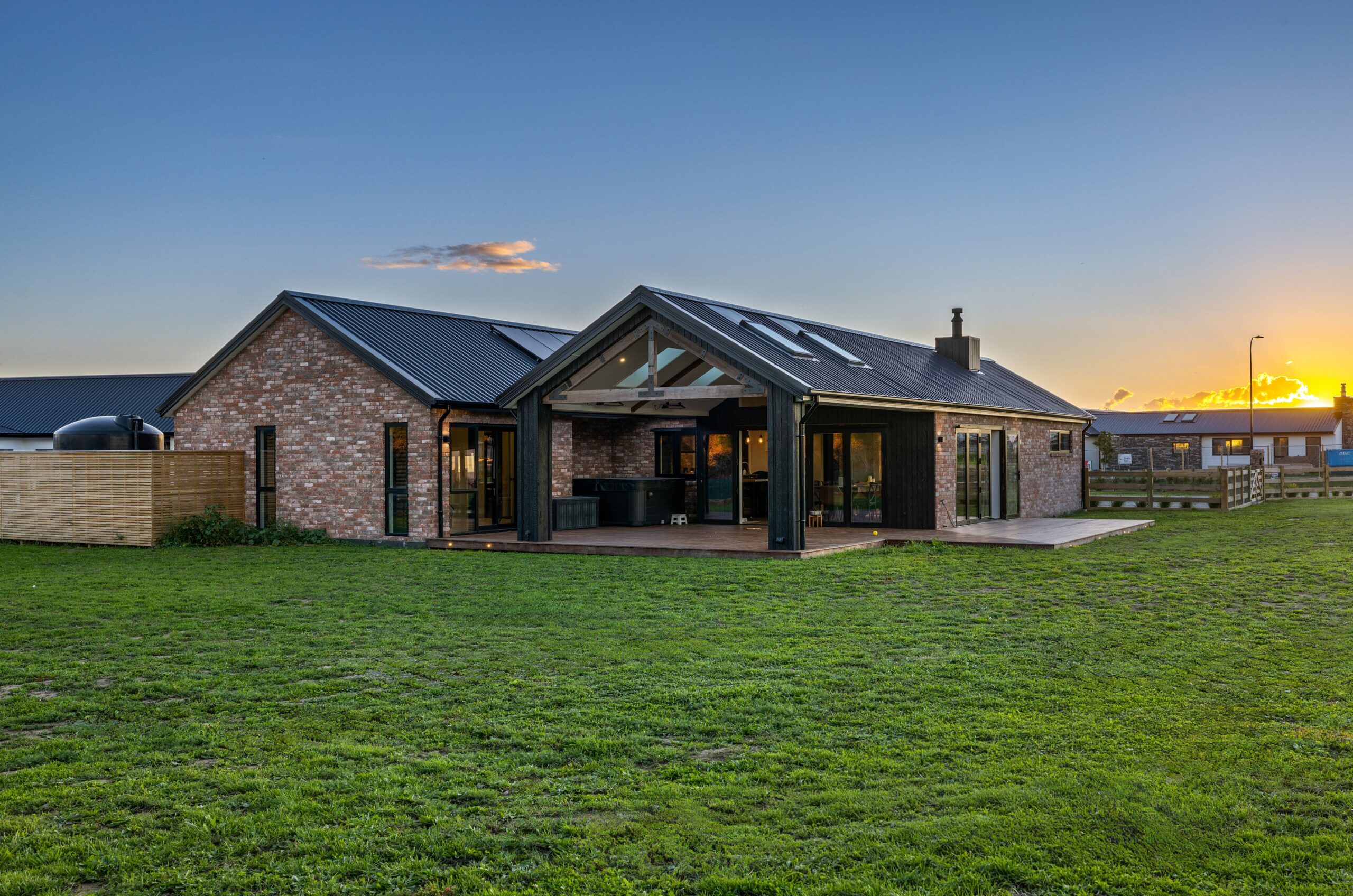 Rear elevation of a Hallmark Homes brick home at golden-hour sunset showing a covered alfresco gable with exposed timber beams and glazing, bi-fold doors opening to a timber deck, multiple roof skylights, and a wide green lawn under a warm orange and blue sky.