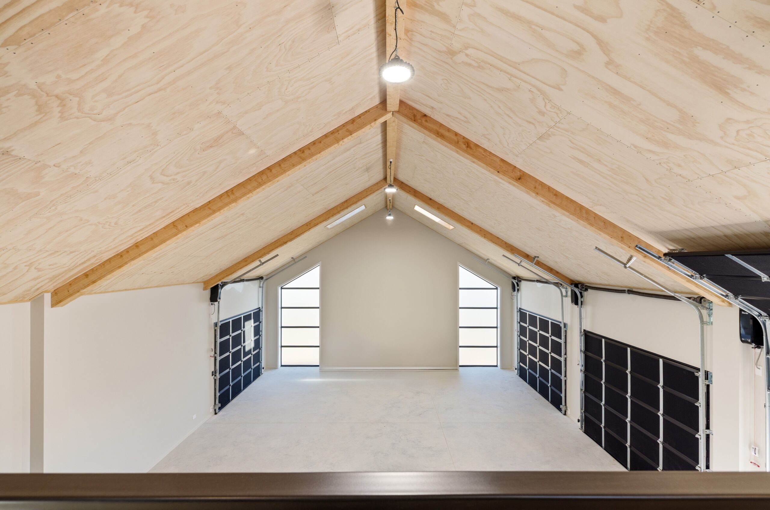 Interior view of a Hallmark Homes triple garage from the mezzanine, showing a symmetrical plywood vaulted ceiling with timber purlins, two black sectional garage doors with glazed panel inserts, and arched framed windows flooding the concrete floor space with natural light.