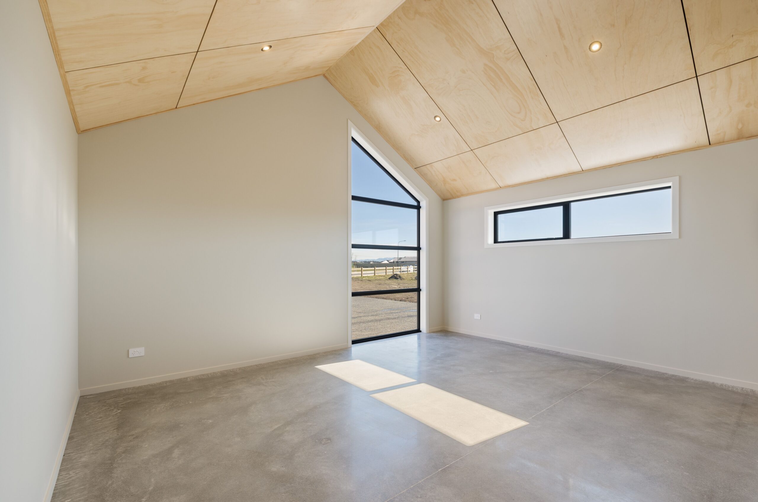 Light-filled flexible room in a Hallmark Homes lifestyle build with a plywood vaulted ceiling, polished concrete floor, recessed downlights, a full-height black-framed glazed tilt-up door opening to the exterior, and a horizontal black-framed clerestory window to the side.