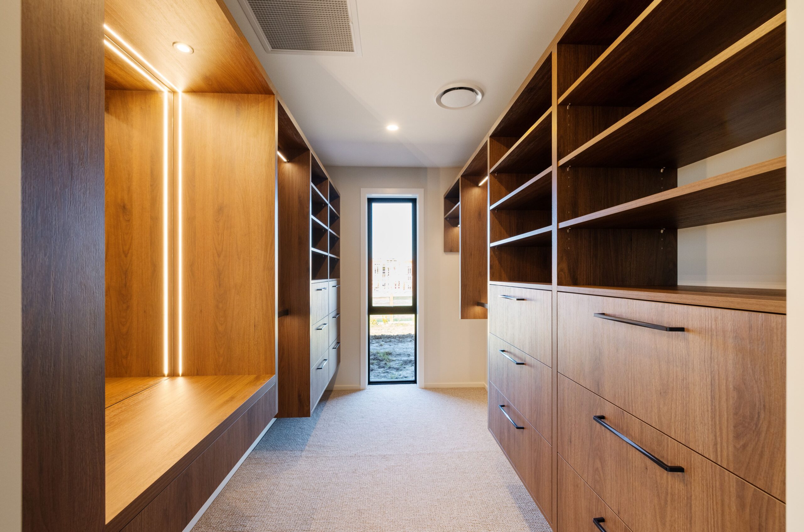 Generous walk-in wardrobe with full-height custom timber joinery on both sides, open shelving, deep drawers with black bar handles, integrated LED strip lighting illuminating the hanging and shelf sections, a carpeted floor, and a narrow black-framed window at the far end.