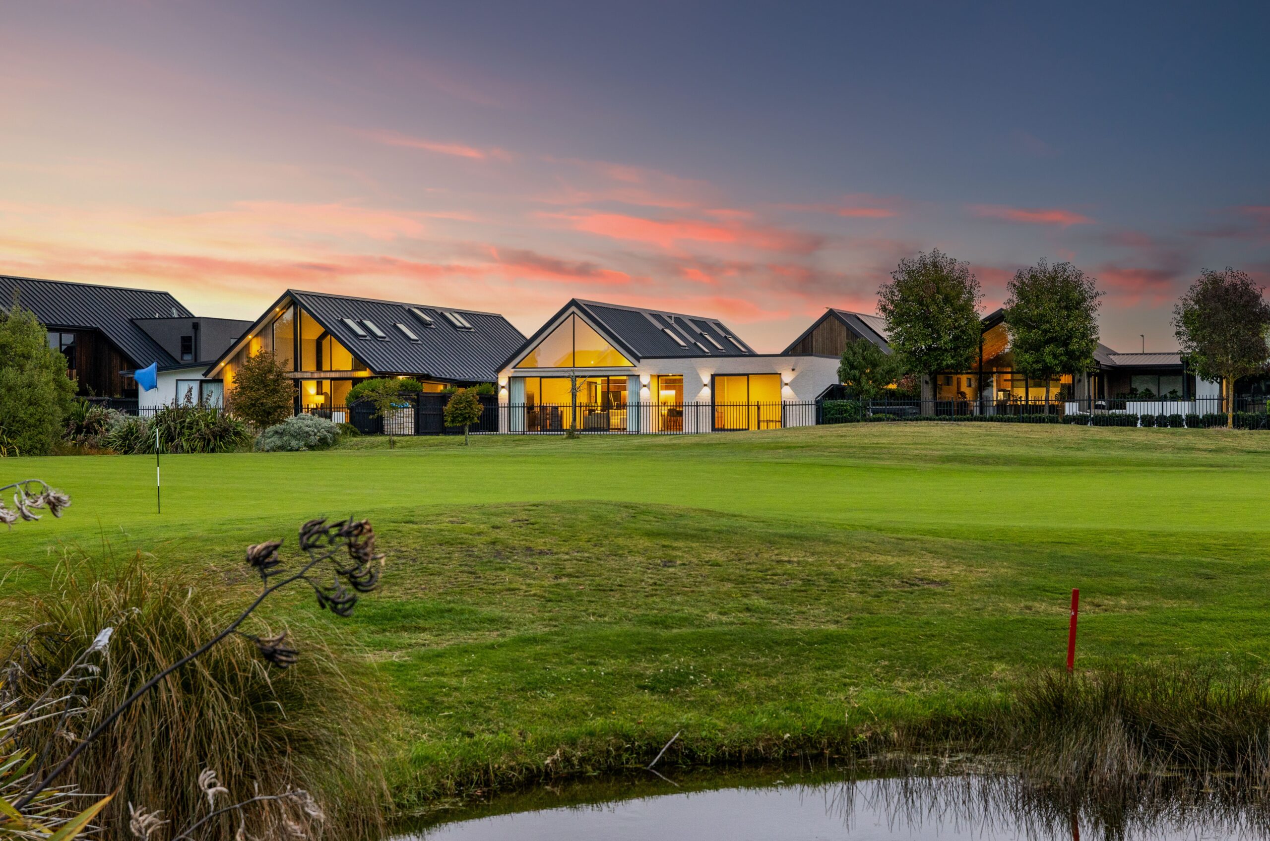 Row of gable-roofed Hallmark Homes overlooking a golf course pond at dusk, pink sunset sky, black metal fencing along the fairway. Hallmark Homes golf course community, Marshlands, Christchurch.