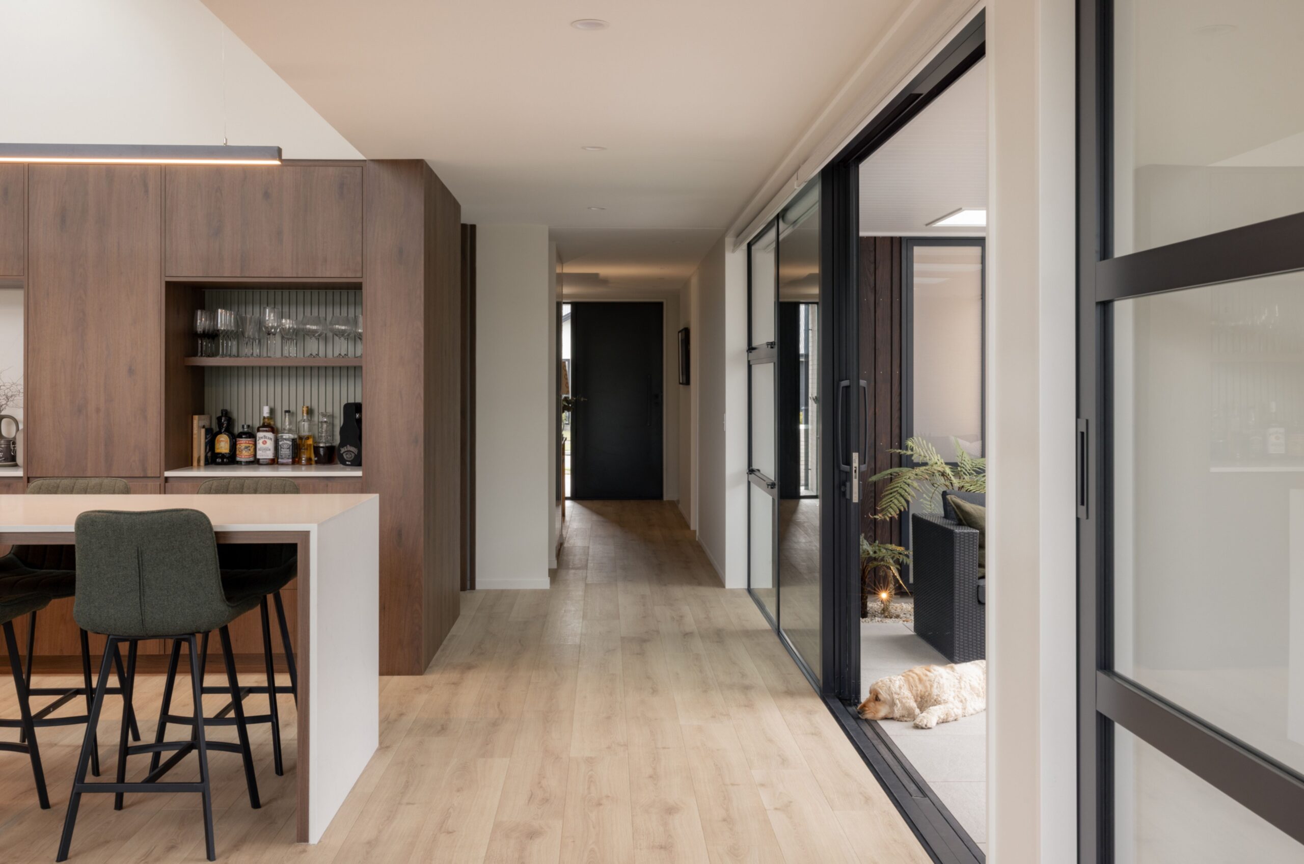Kitchen hallway with dark walnut cabinetry, built-in bar nook with glassware and spirits, black sliding glass doors leading to a courtyard. Hallmark Homes kitchen joinery, Marshlands, Christchurch.