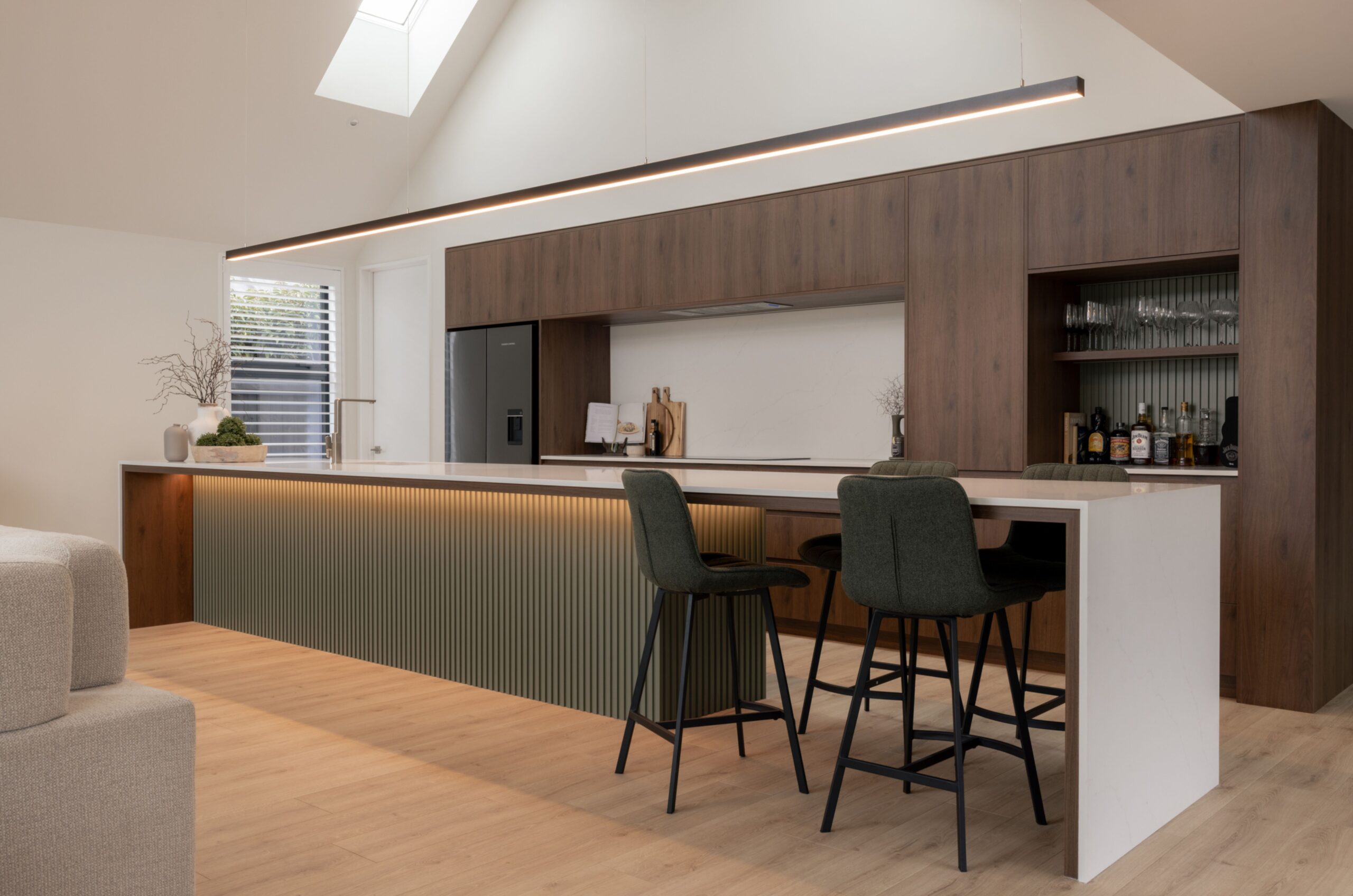 Vaulted kitchen with dark walnut cabinetry, sage green fluted island panelling, black bar stools, linear pendant light, and skylights. Hallmark Homes kitchen design, Marshlands, Christchurch.