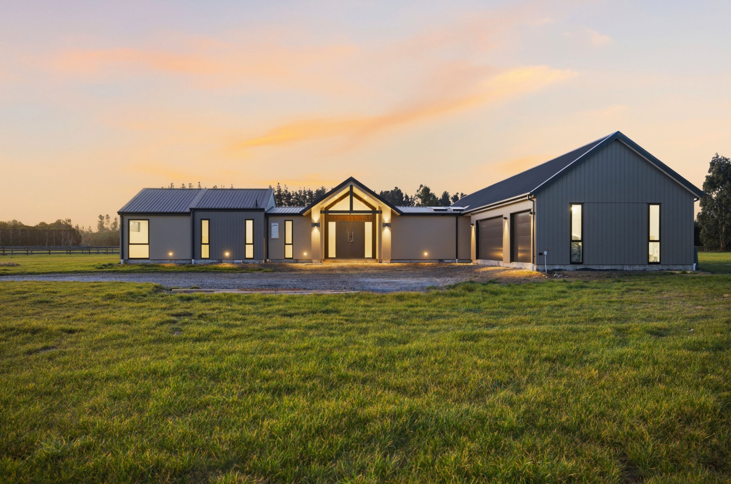 Wanaka plan, Hallmark Homes West Melton. Dusk view of the triple garage wing and gravel driveway lit against a pink sky.