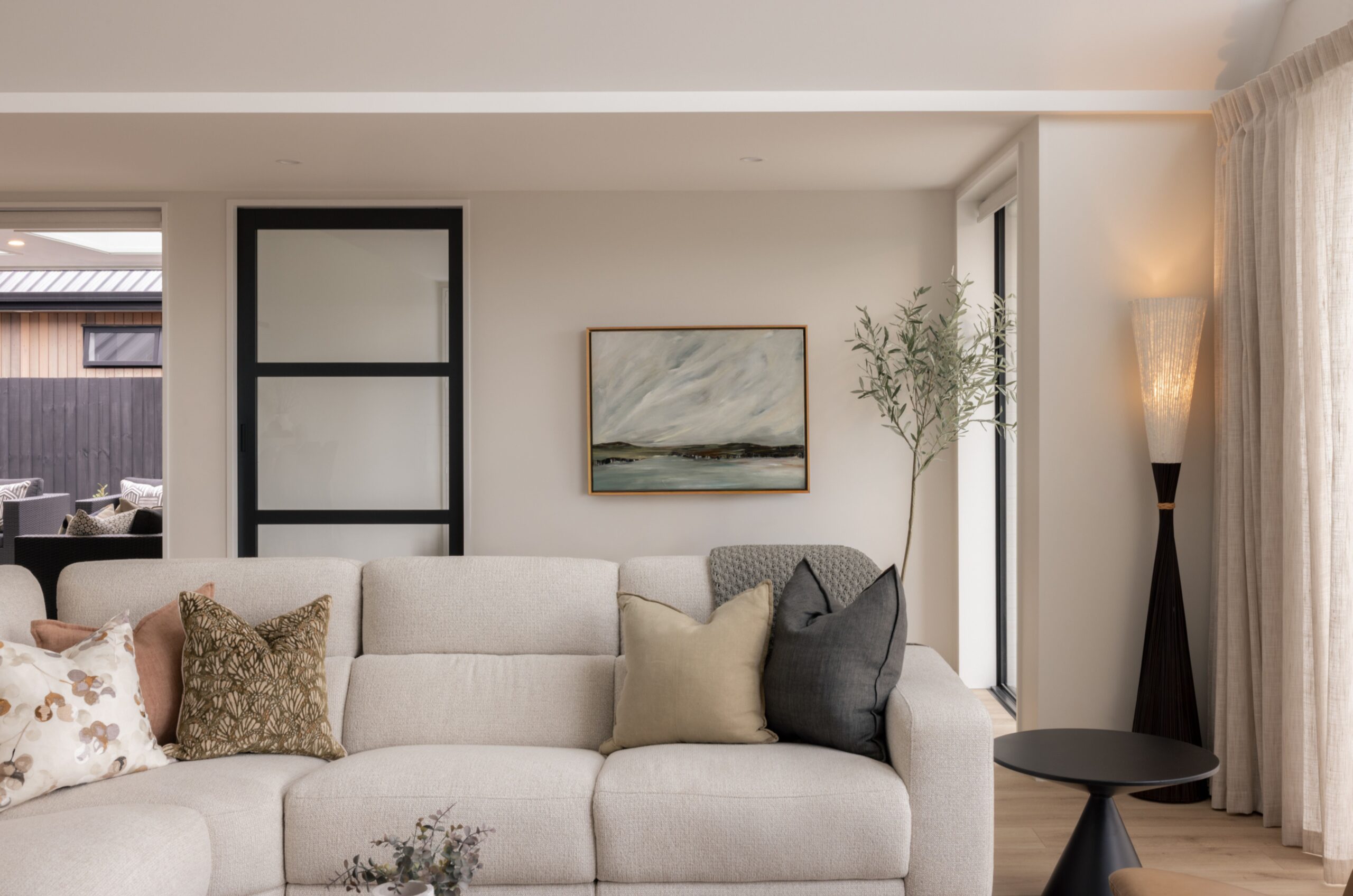 Living room corner with cream sofa, mixed cushions, framed coastal artwork, olive tree, and black floor lamp beside sheer curtained windows. Hallmark Homes lounge styling, Marshlands, Christchurch.