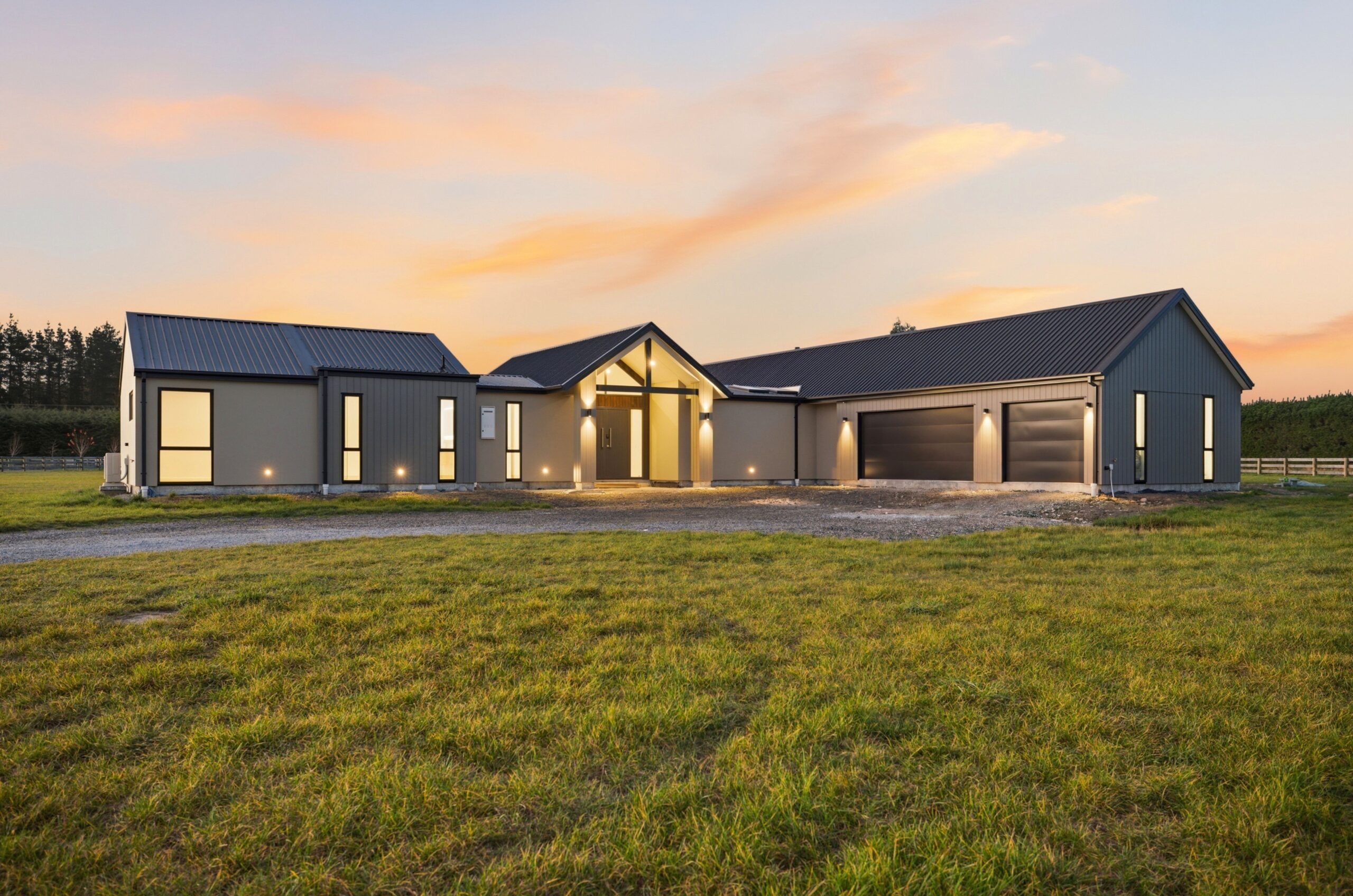 Wanaka plan by Hallmark Homes, West Melton. Front elevation at dusk with charcoal and grey cladding, gabled rooflines, and an illuminated timber entry.