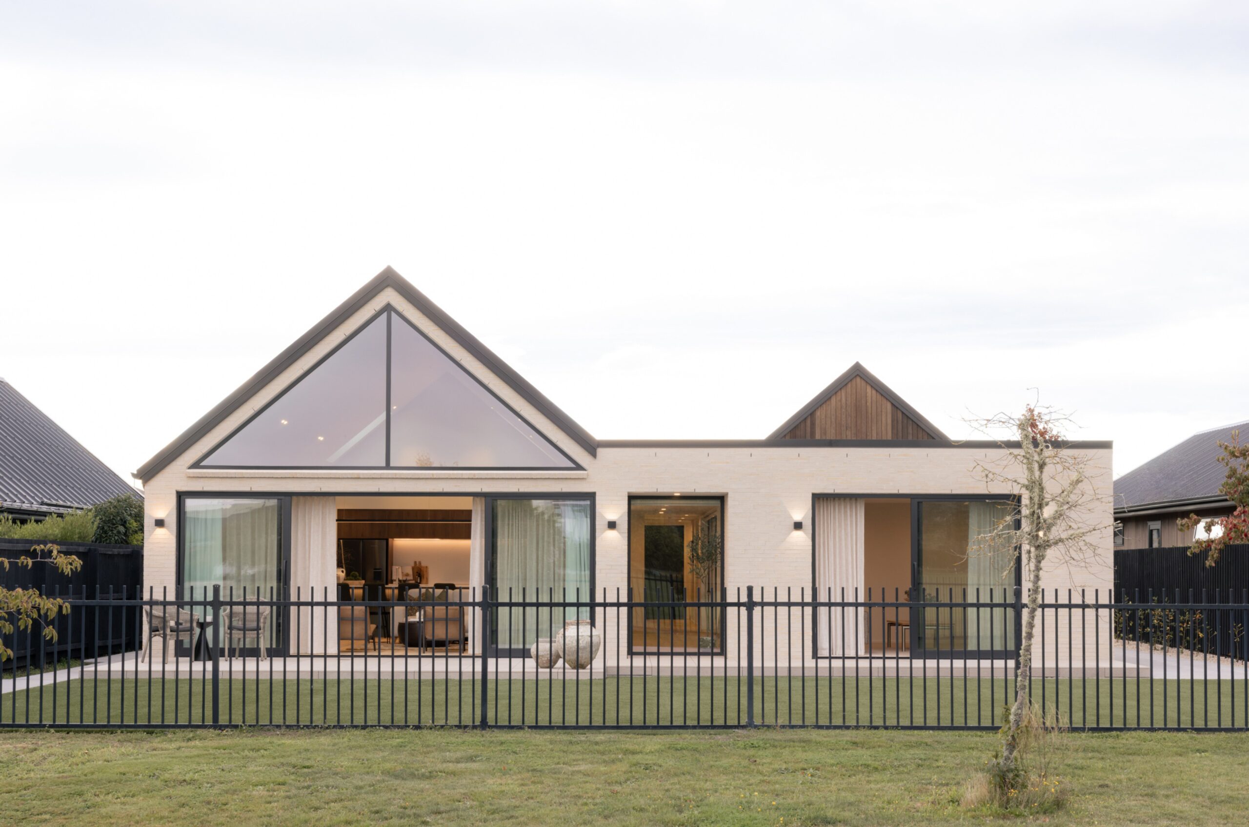 Front exterior of a brick Hallmark Homes build with a large triangular gable window, timber gable accent, black aluminium joinery, and black picket fence. Hallmark Homes facade design, Marshlands, Christchurch.