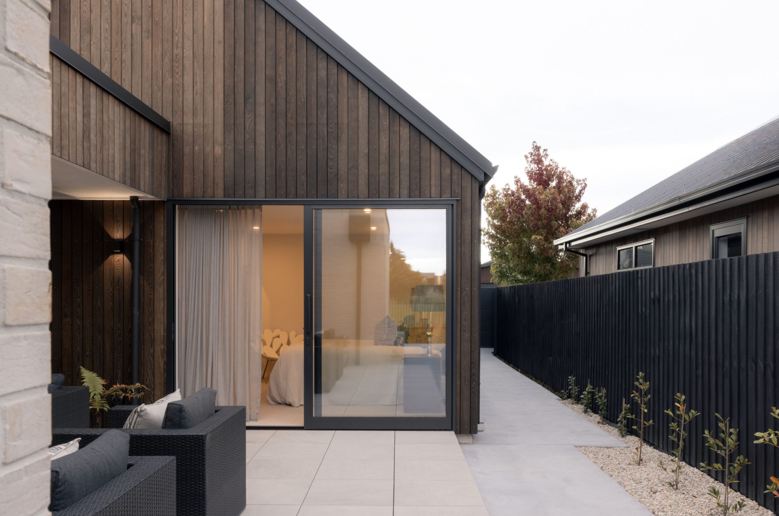 Modern black timber-clad home exterior with gable roof, black aluminium sliding doors opening to a bedroom, outdoor lounge seating, and a black slatted privacy fence. Hallmark Homes new build, Marshlands, Christchurch.