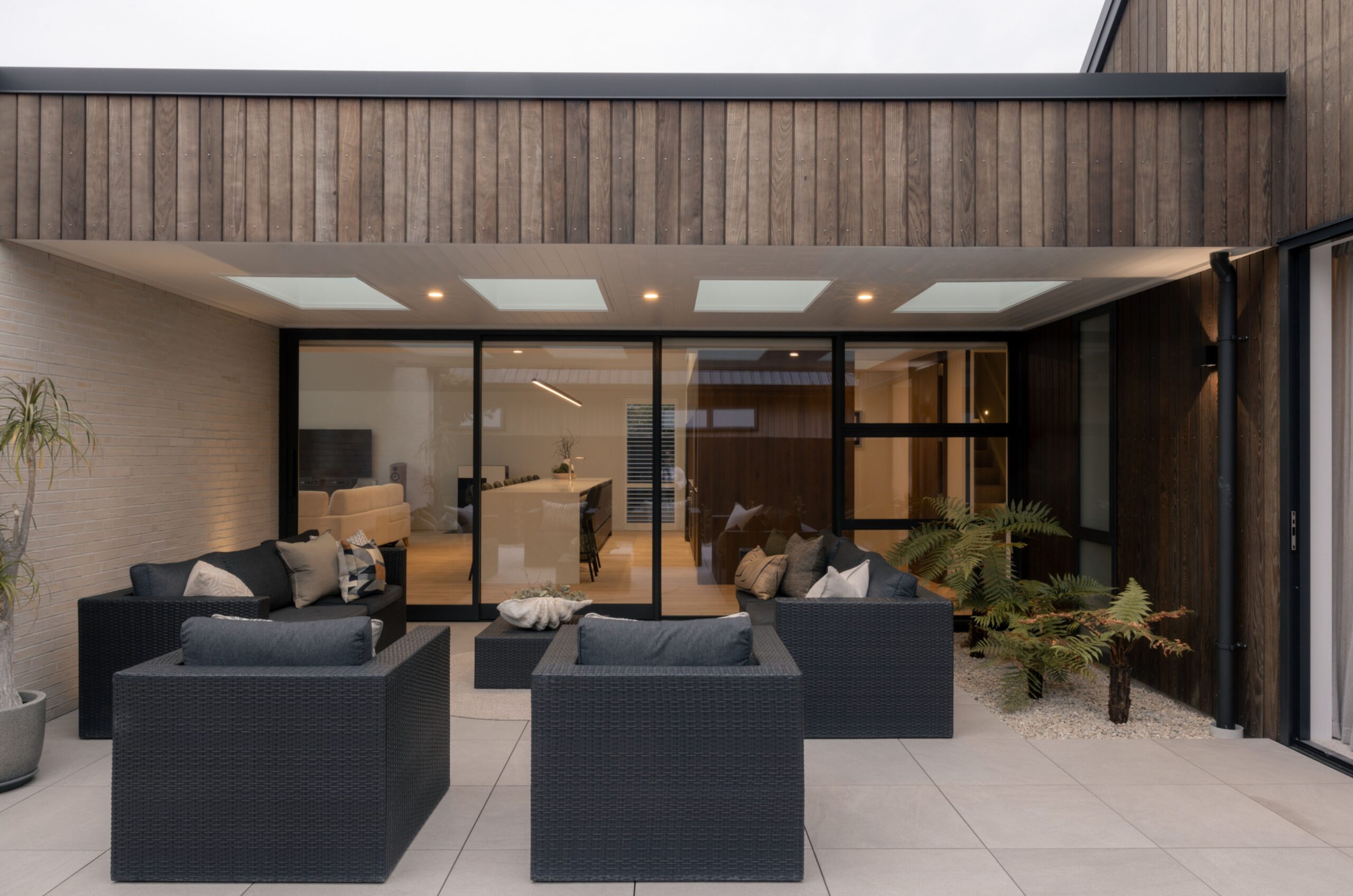 Covered outdoor patio with skylights and black wicker sofas, opening through glass sliding doors to an open-plan kitchen and living area with brick feature wall. Hallmark Homes indoor-outdoor living, Marshlands, Christchurch.