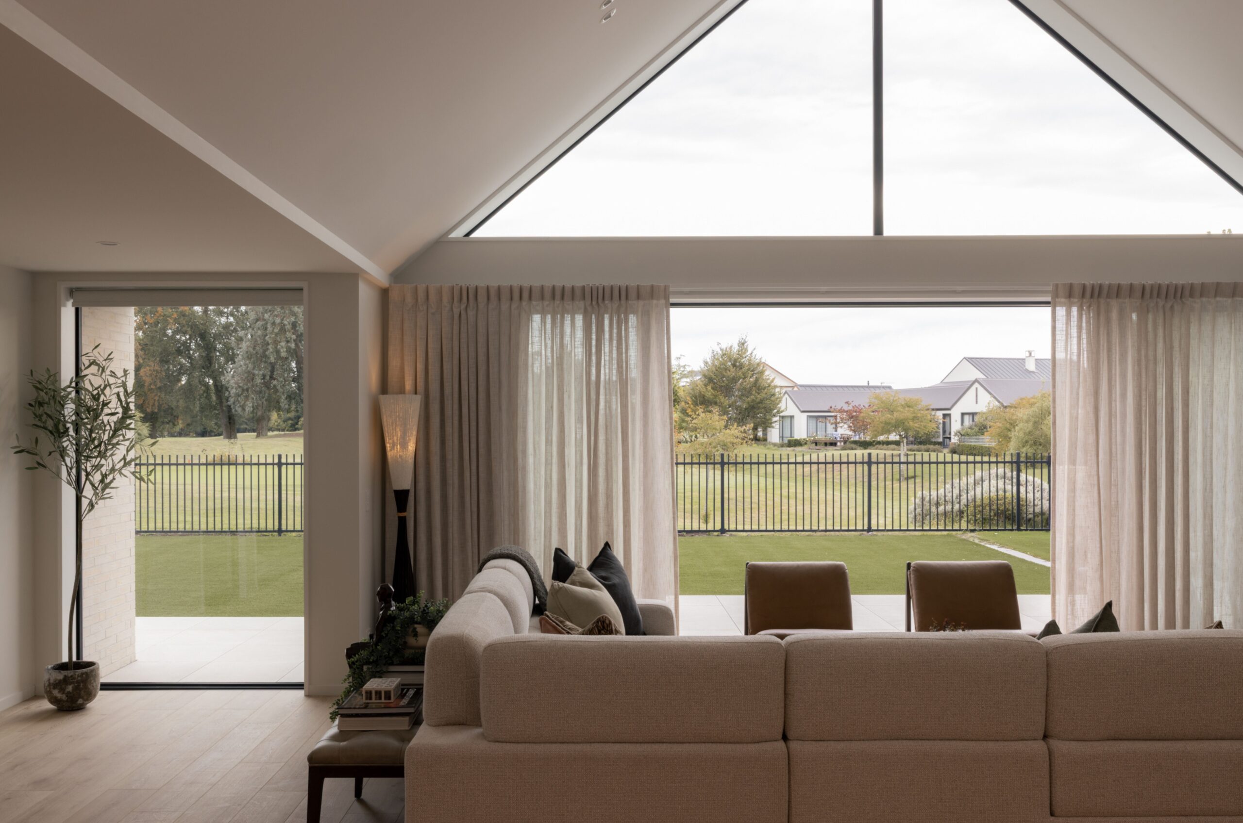Open-plan living room with vaulted gable window, sheer linen curtains, beige sectional sofa, and leather accent chairs overlooking a golf course fairway. Hallmark Homes lounge interior, Marshlands, Christchurch.