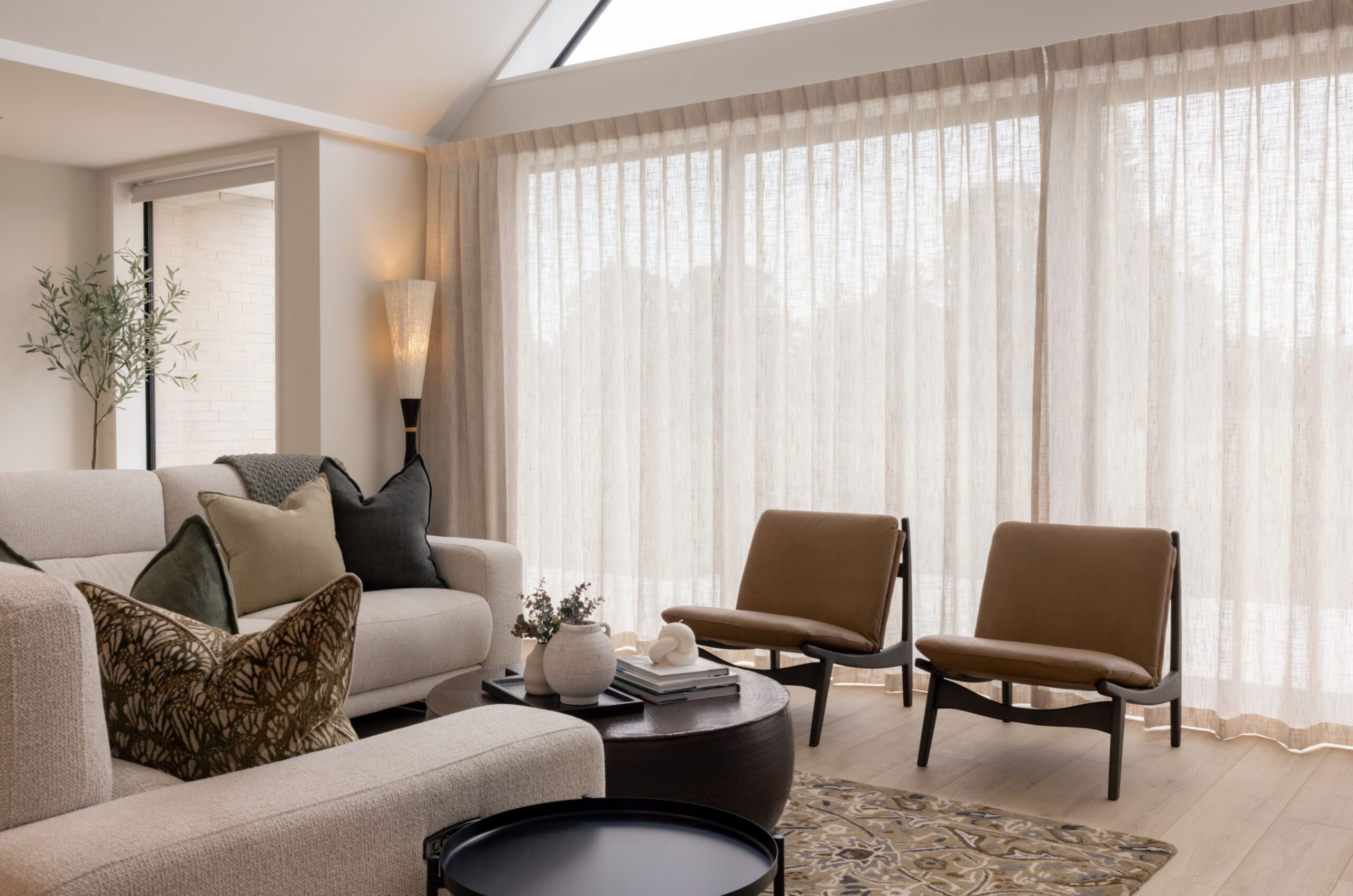 Living room seating area with sheer curtains, tan leather lounge chairs, patterned rug, and a round dark wood coffee table. Hallmark Homes furnished show home, Marshlands, Christchurch.