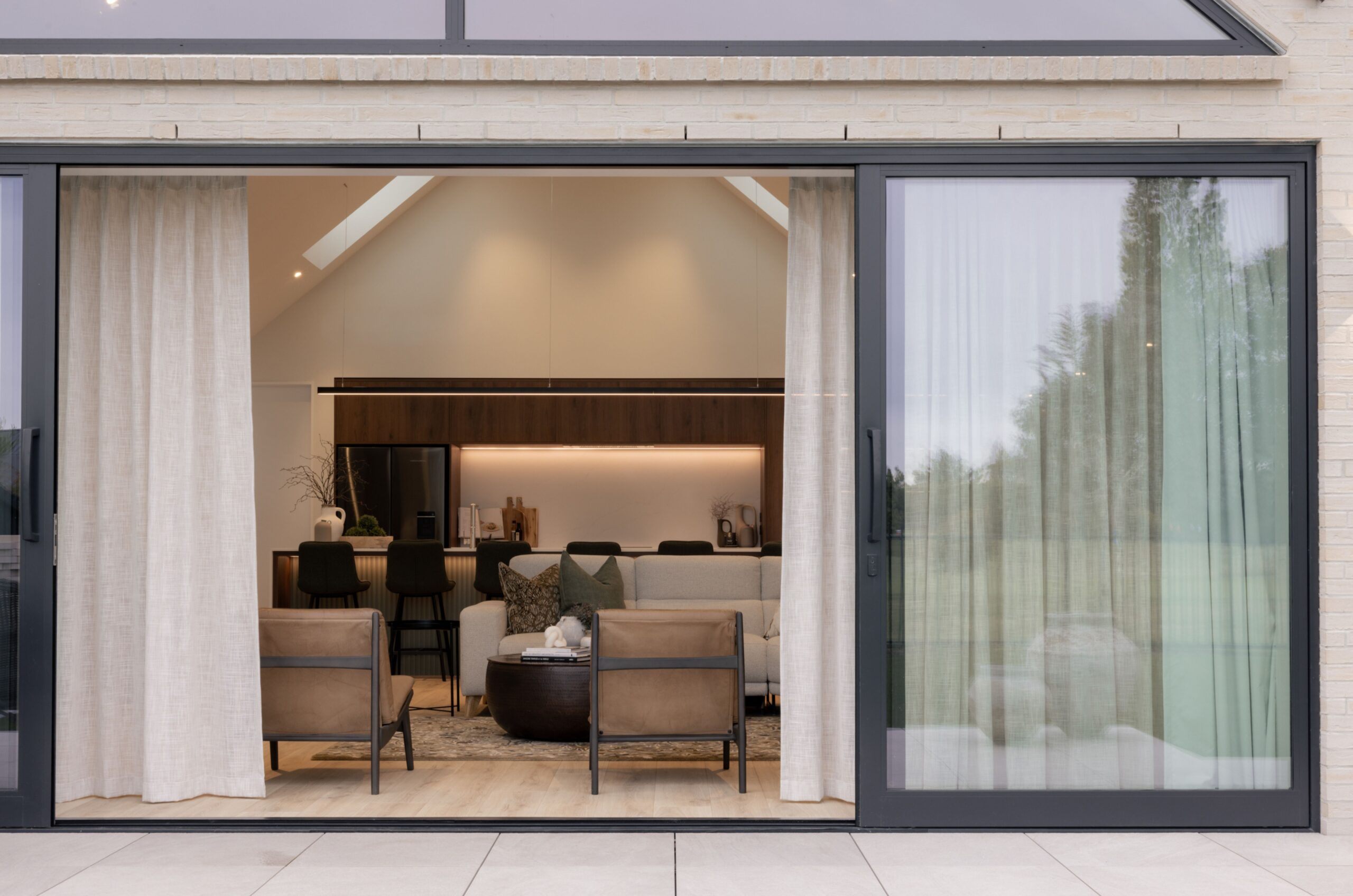 Close-up of open black-framed sliding doors with sheer curtains revealing a vaulted kitchen and living room, brick exterior cladding. Hallmark Homes indoor-outdoor flow, Marshlands, Christchurch.